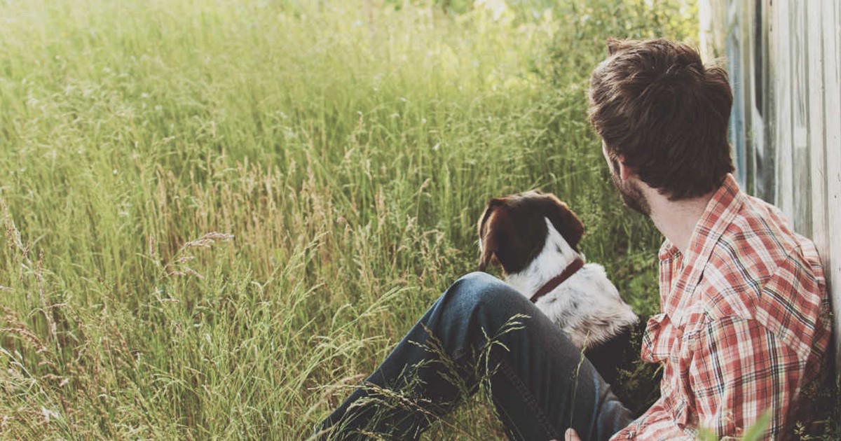 young man sitting with his dog in long grass against a wall